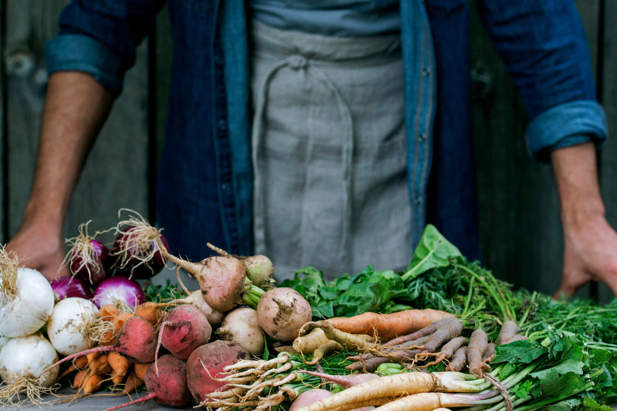 Farmer in a denim shirt and apron behind a table of freshly harvested root vegetables and greens