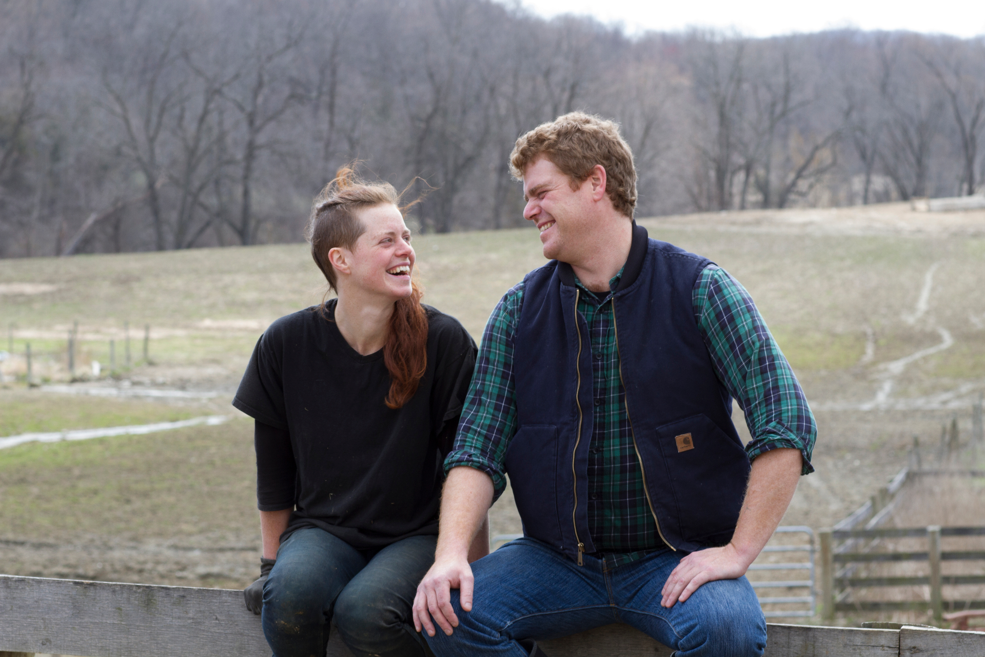 Two farmers laughing together while sitting on a wooden fence at their farm